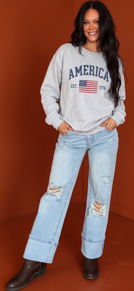 Crown N Thread grey crewneck sweatshirt with distressed America 1776 flag print worn by a smiling woman in jeans.
