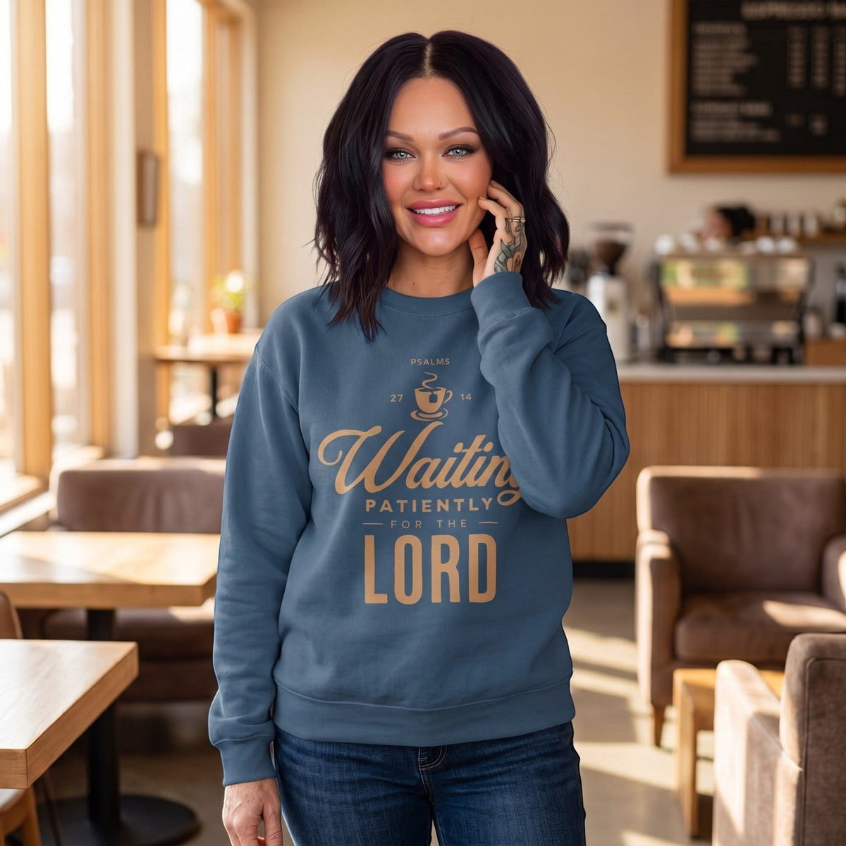 A woman wearing a Crown N Thread blue crewneck sweatshirt with Waiting Patiently for the Lord text in a cafe.