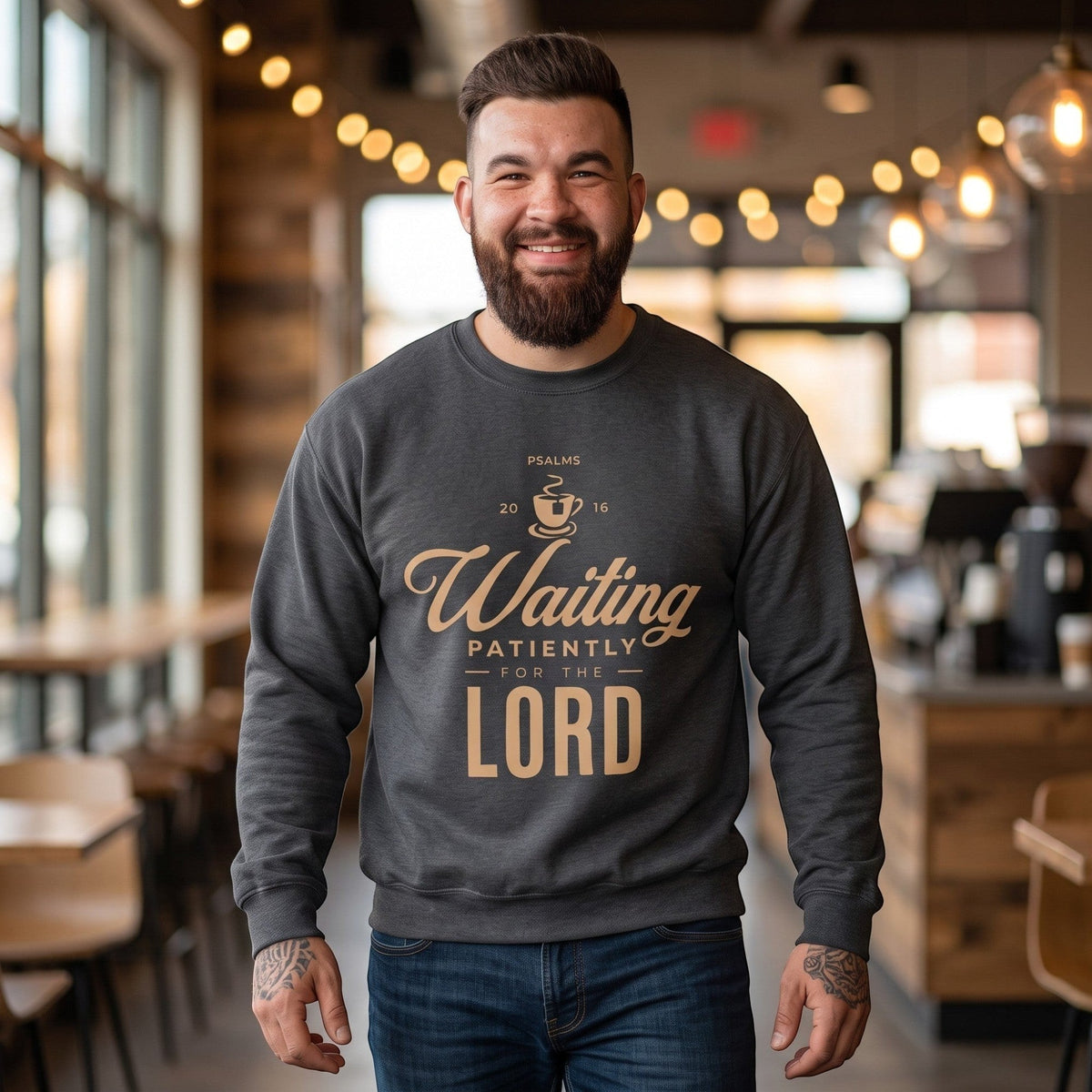A man wearing a Crown N Thread charcoal grey crewneck sweatshirt with Waiting Patiently for the Lord text in a cafe.